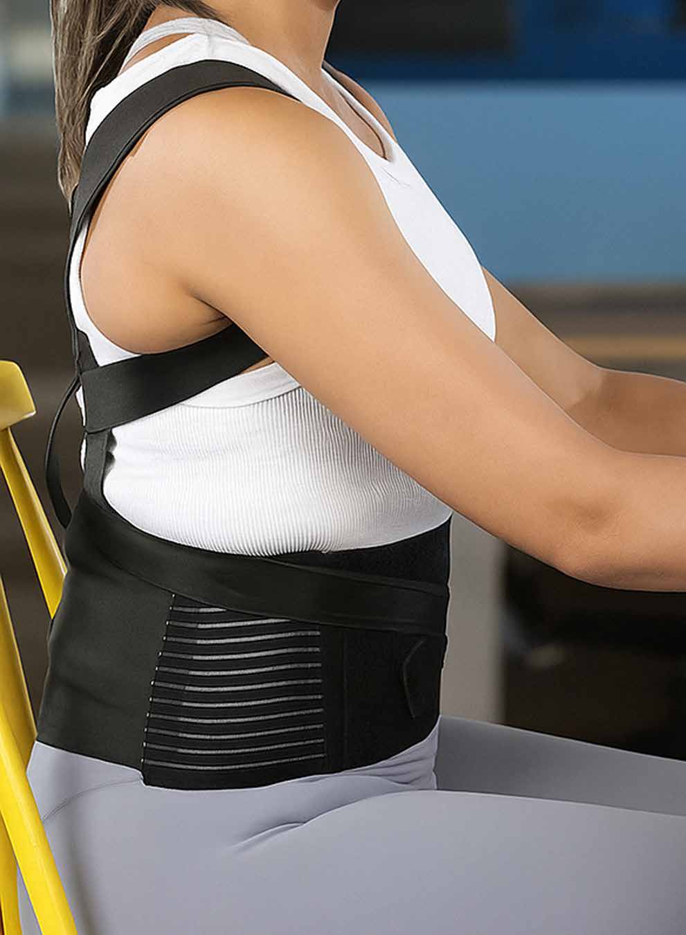 Model wearing the black posture corrector over a white tank top while seated, straps crossing the upper back and shoulders.